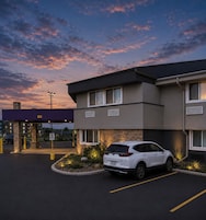 A modern hotel with a white SUV parked in the driveway, a no parking sign, and a sunset sky.