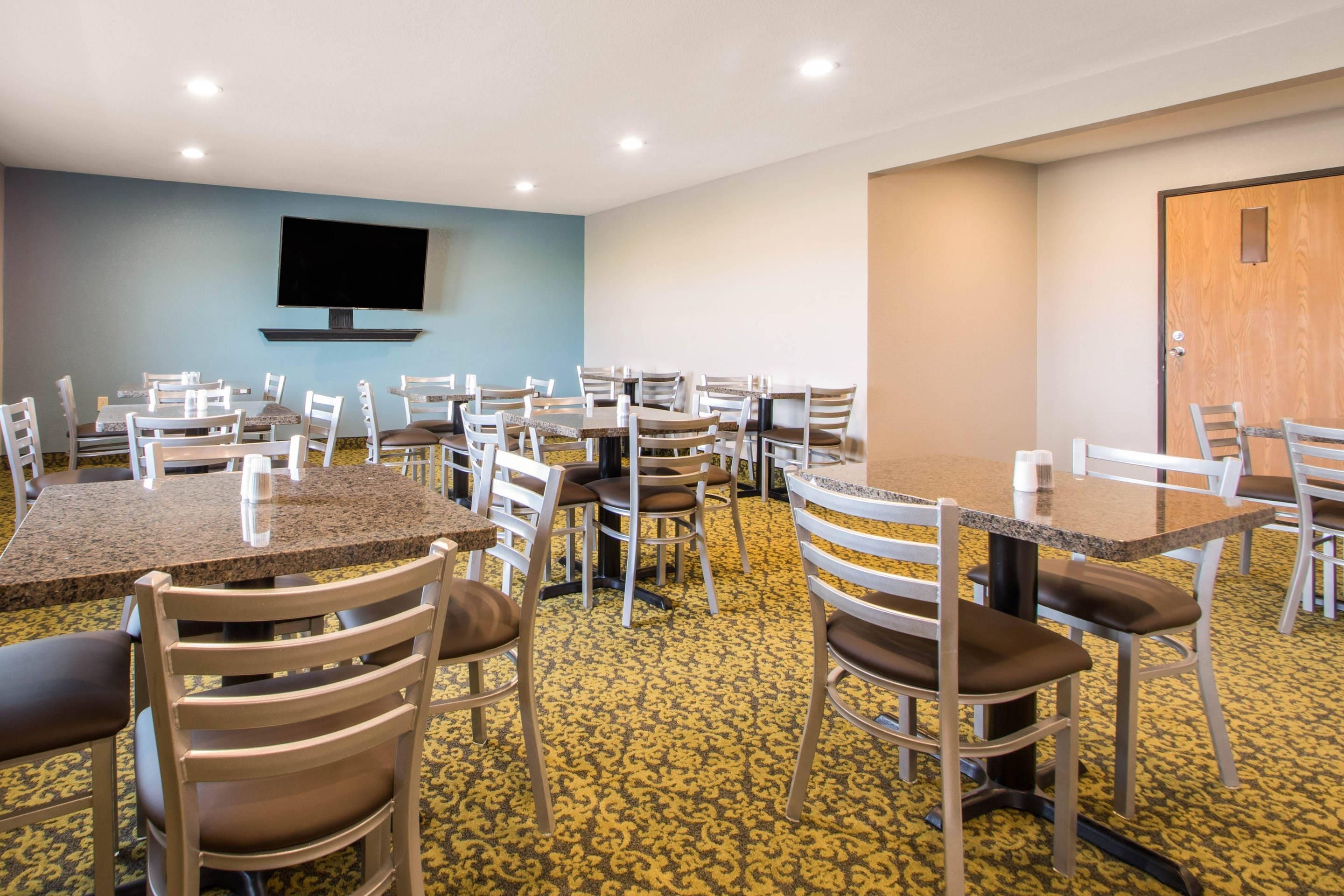A hotel dining area with multiple tables and chairs, a flat-screen TV mounted on the wall, and a patterned carpet.