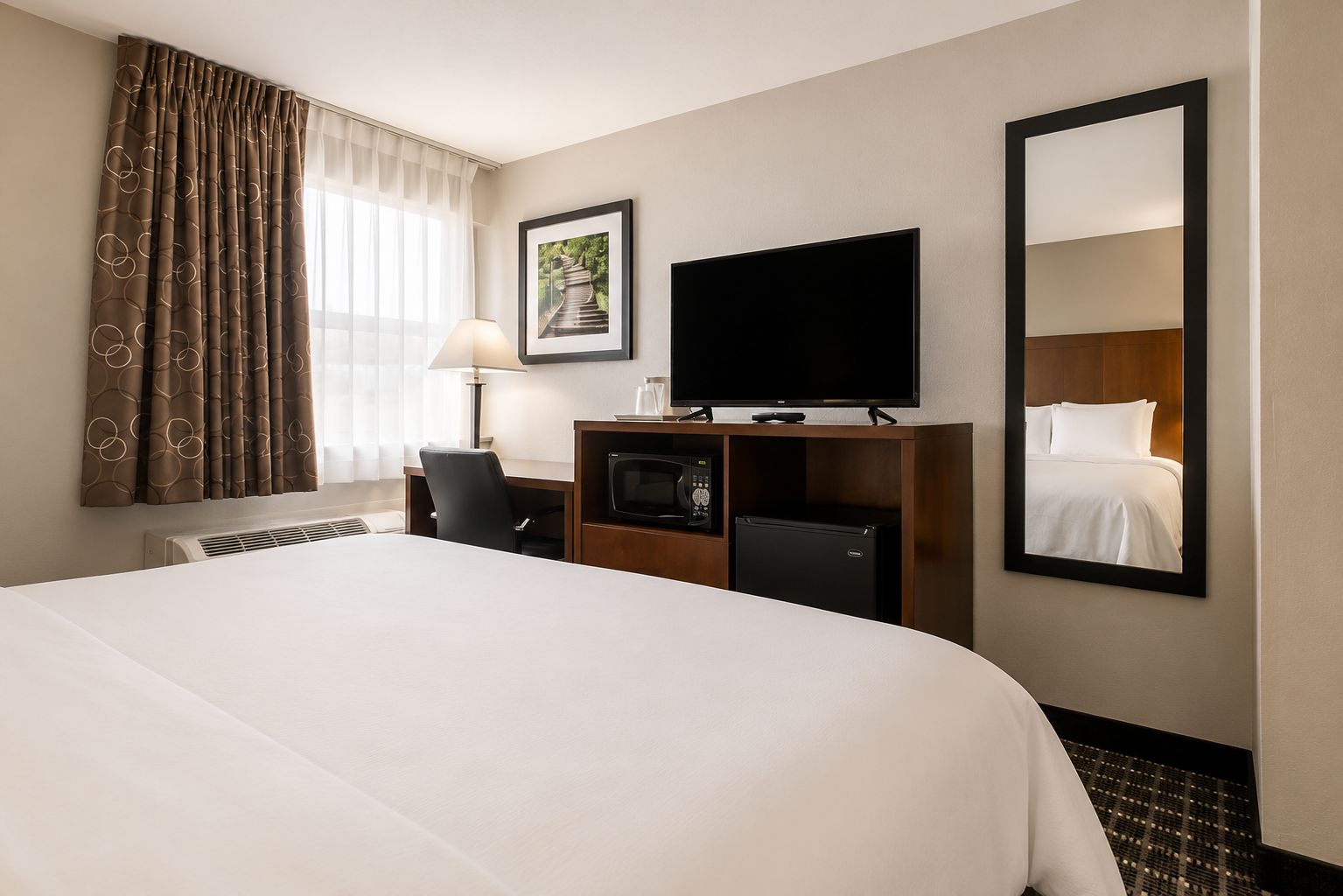 Une chambre d’hôtel dotée d’un grand lit, d’un téléviseur à écran plat, d’un bureau avec un ordinateur, d’un réfrigérateur et d’un miroir qui reflète le lit.