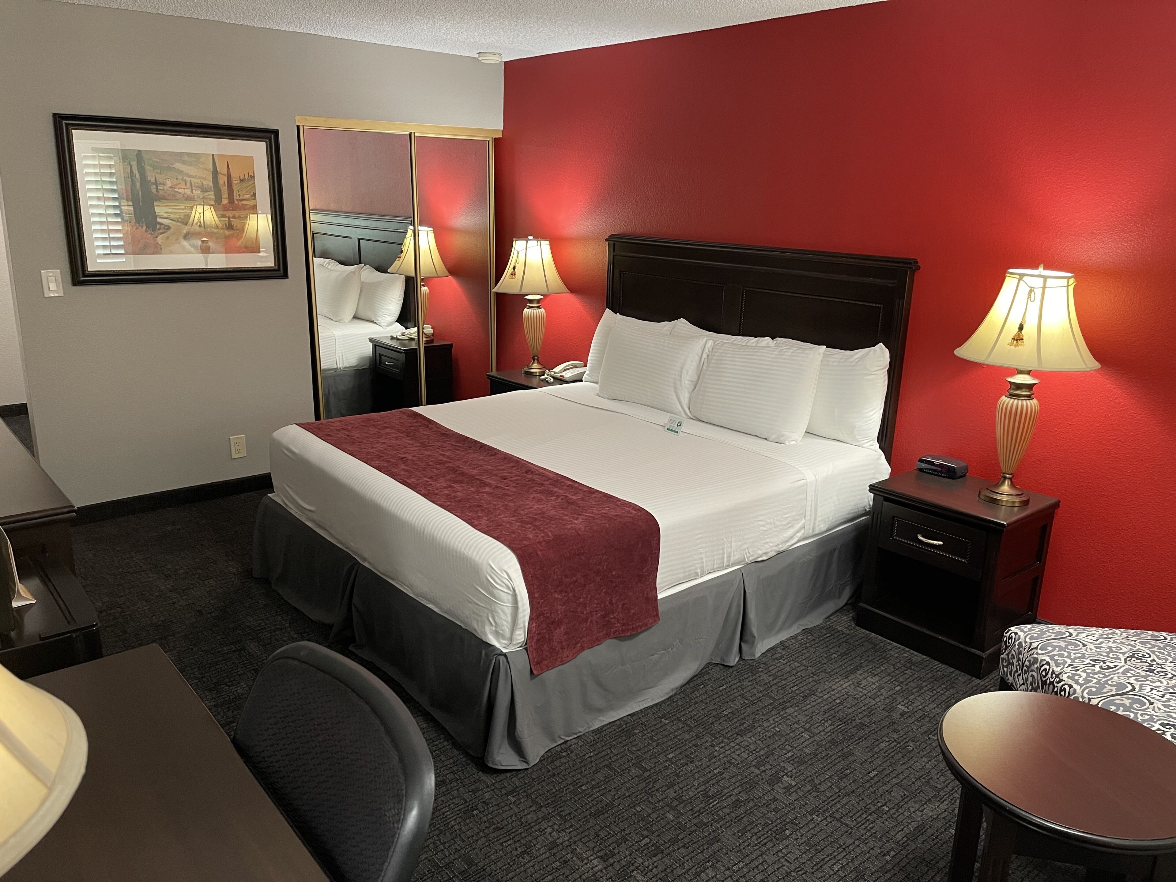Habitación de hotel con una cama grande, dos mesitas de noche con lámparas, un escritorio con silla, un espejo y un cuadro en la pared.