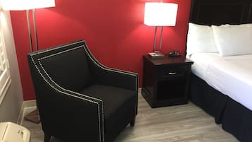 Une chambre d’hôtel avec un fauteuil noir à revêtement en tissu, une table de chevet en bois avec une lampe, un lit avec du linge de lit blanc et un mur rouge.