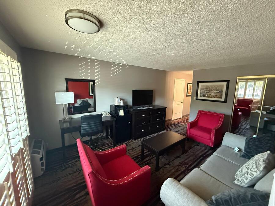 Habitación de hotel con sala de estar que incluye un sofá, un sillón rojo, una mesa de centro, un televisor, un escritorio con lámpara y un espejo.