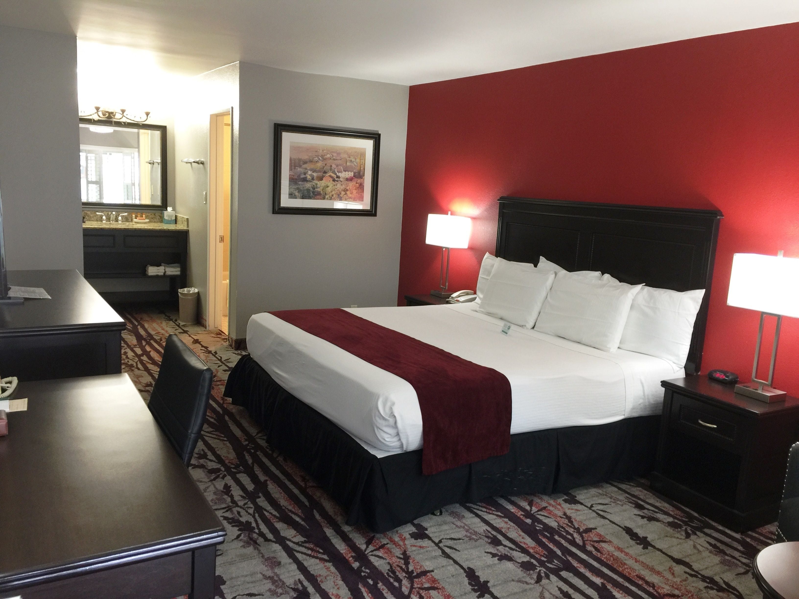 A hotel room with a large bed, two bedside lamps, a desk, a chair, a mirror, and a bathroom visible through an open door.