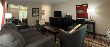 A living room with a sofa, armchair, and a small table. There is a television, a microwave, and a lamp on a desk.