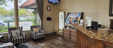Un área de recepción con un sillón a cuadros, un mostrador de madera con una exhibición de folletos, un reloj y un televisor montado en la pared.