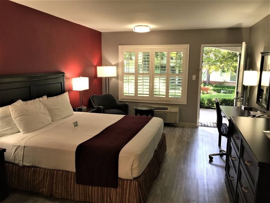 Quarto de hotel com uma cama grande, duas luminárias de cabeceira, uma escrivaninha com cadeira, um penteado e uma janela com persianas.