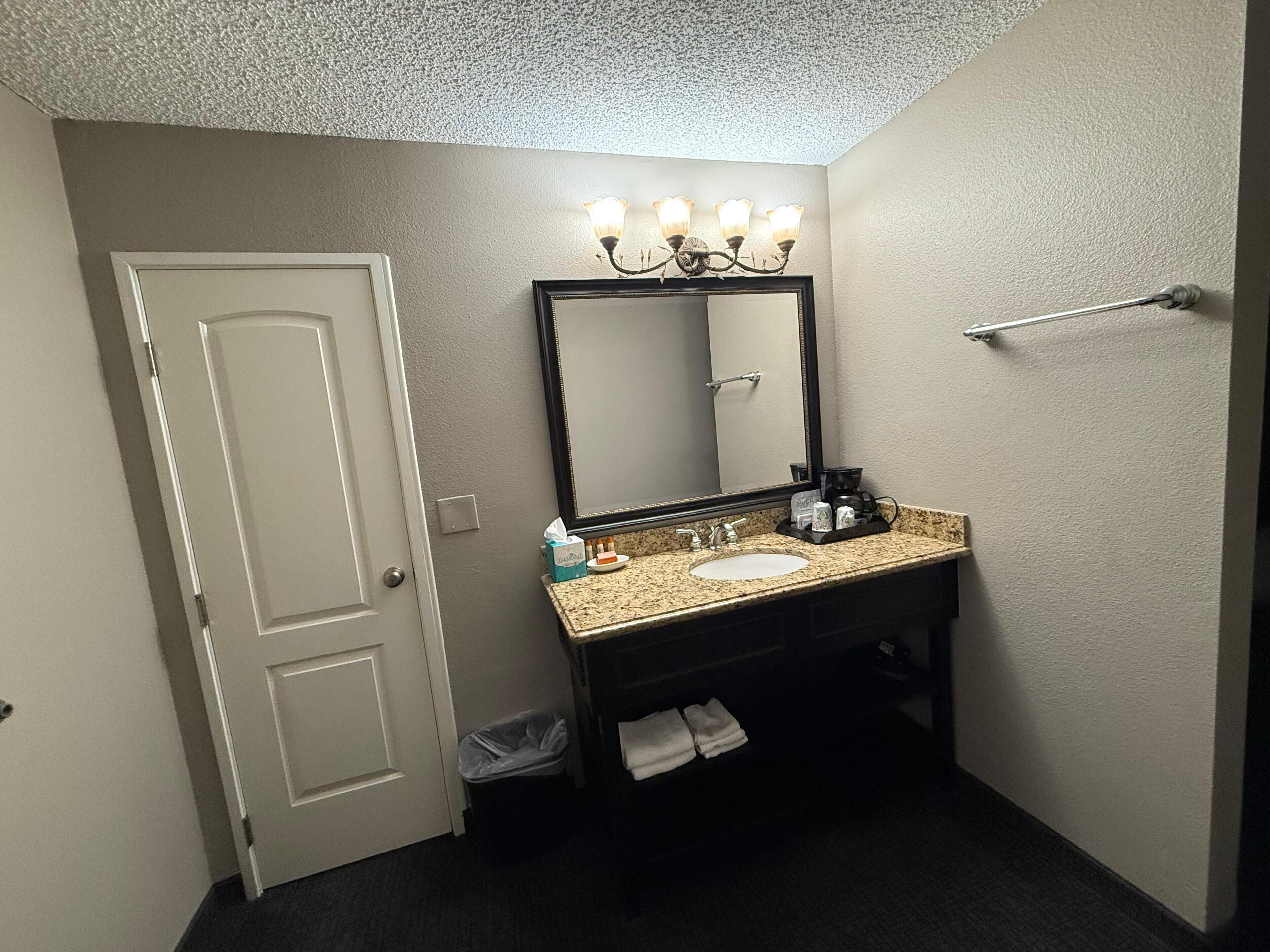 A bathroom with a white door, a vanity with a granite countertop, a mirror with lights, a towel rack, and a trash bin.