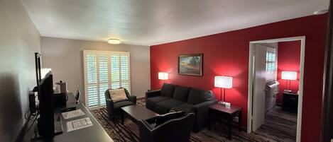 A hotel room with a dark-colored sofa, a small table, a desk with a computer, and two lamps. There is a window with blinds and a door leading to another room.