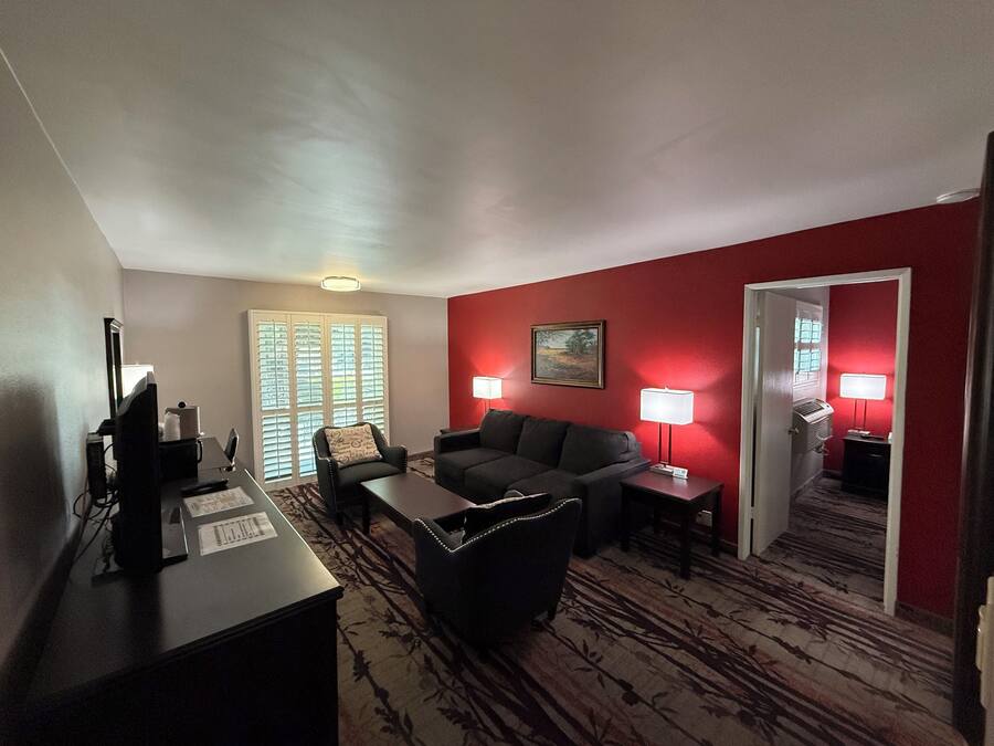 A hotel room with a dark-colored sofa, a small table, a desk with a computer, and two lamps. There is a window with blinds and a door leading to another room.