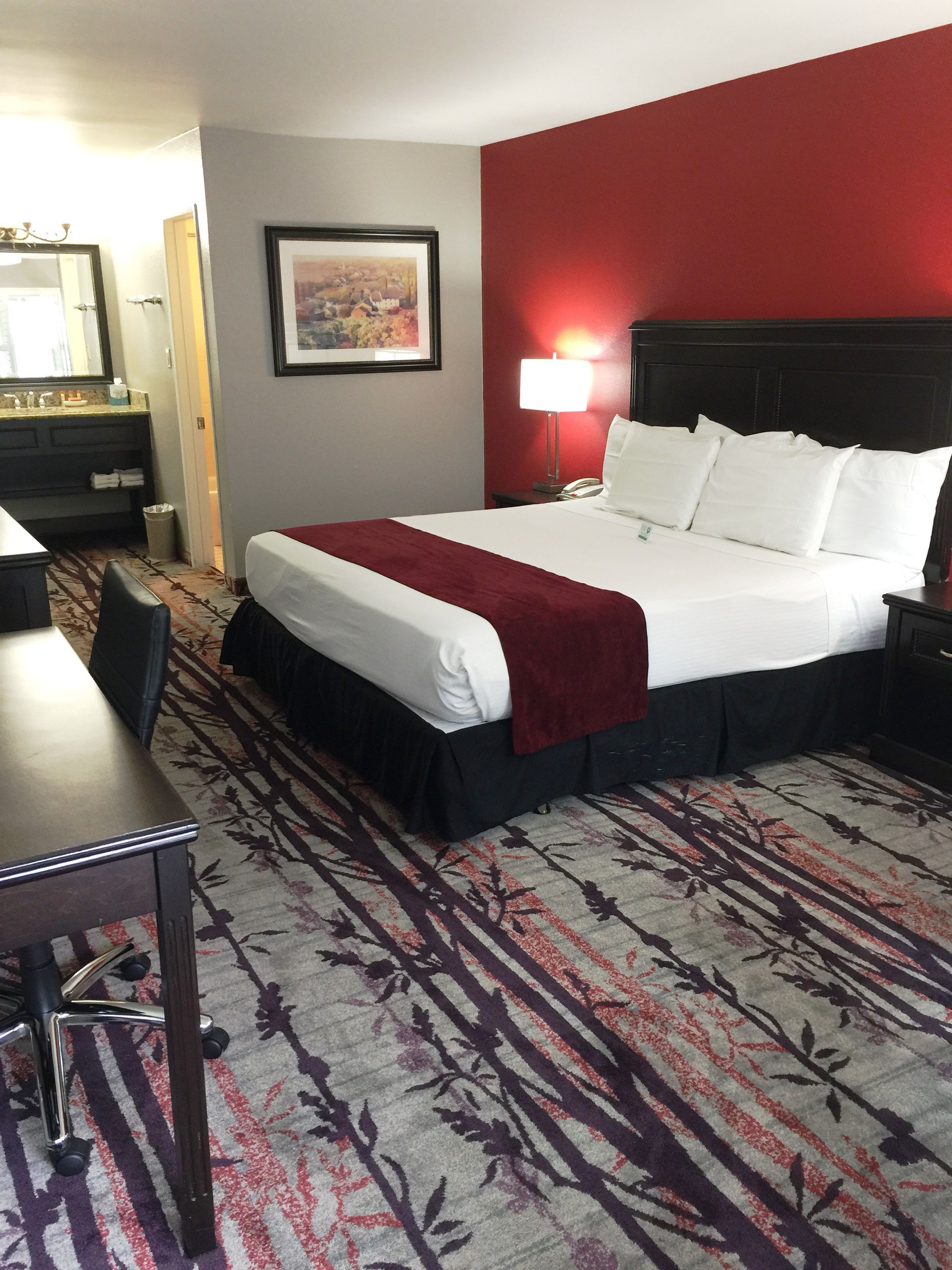 Habitación de hotel con una cama grande, un escritorio, una silla, una lámpara y un cuadro en la pared.