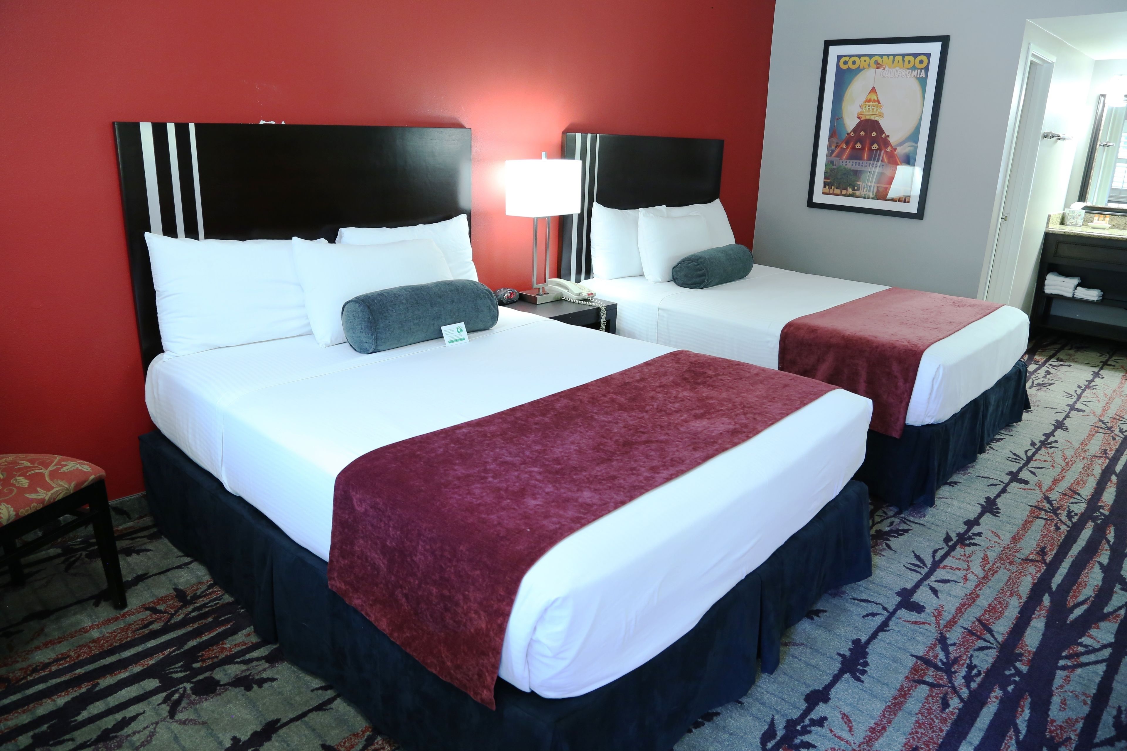 Ein Hotelzimmer mit zwei Betten, einer roten Wand, einem gerahmten Bild, einem Nachttisch und einem gemusterten Teppich.