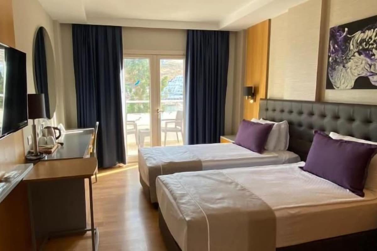 A hotel room with two beds, a desk, a TV, and a balcony with curtains.
