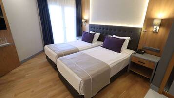 A hotel room with two beds, a wooden floor, a large window with curtains, and a nightstand with a phone.