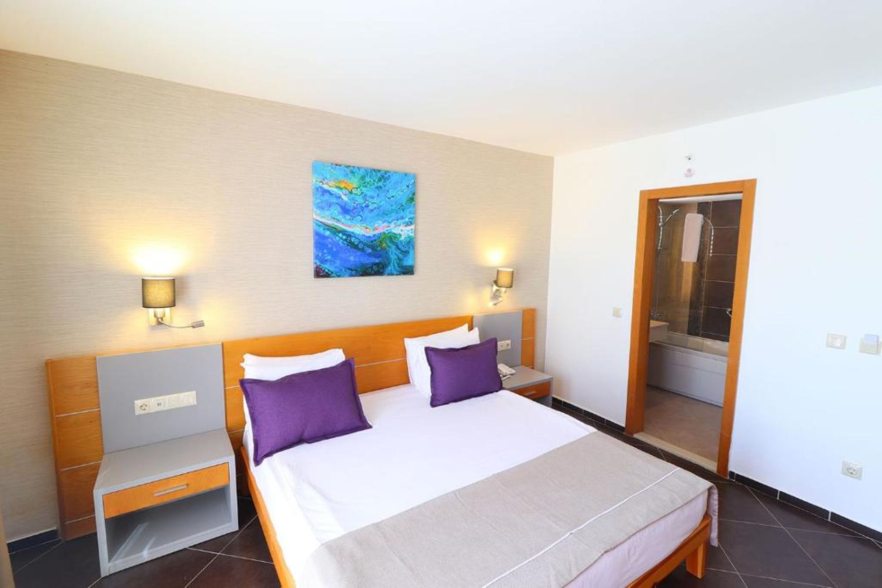 A hotel room with a large bed, two nightstands, a painting on the wall, and a bathroom door visible through an open doorway.