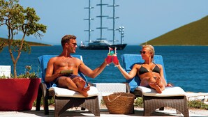 A couple relaxing on sun loungers by the sea with a sailboat in the background. - La Quinta by Wyndham Bodrum (Bodrum)
