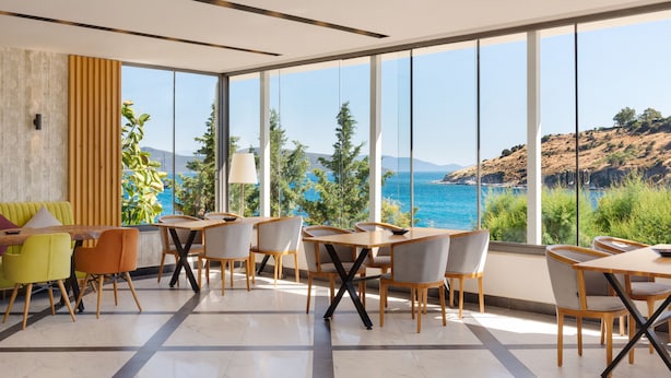A modern dining area with large windows, a view of the sea, and a mix of wooden and upholstered furniture. - La Quinta by Wyndham Bodrum (Bodrum)