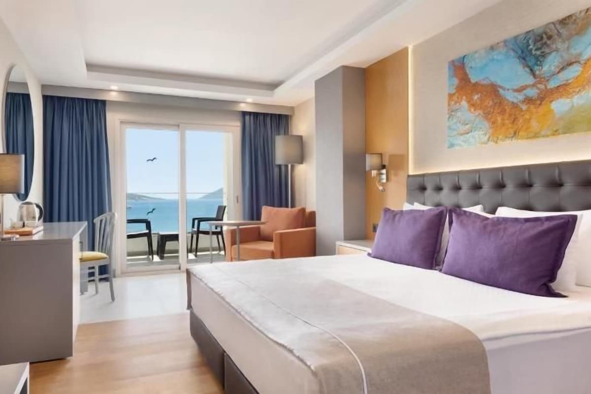 A hotel room with a bed, a desk, a chair, a table, and a view of the sea.