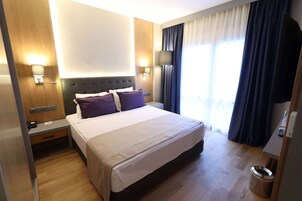 Habitación de hotel con una cama grande, una mesita de noche, una lámpara de pared y una ventana con cortinas.