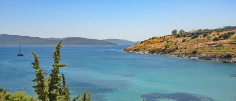 Vista costera con aguas azules cristalinas, un velero y un paisaje montañoso con edificios.