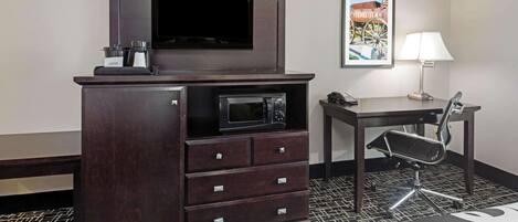 A hotel room with a dark wooden dresser, a flat-screen TV, a microwave, a desk with a chair, a framed picture on the wall, and a bed with a patterned bedspread.