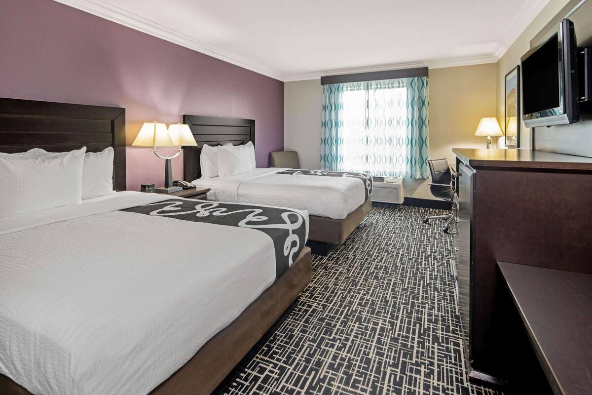 A hotel room with two beds, a desk, a TV, and a window with patterned curtains.