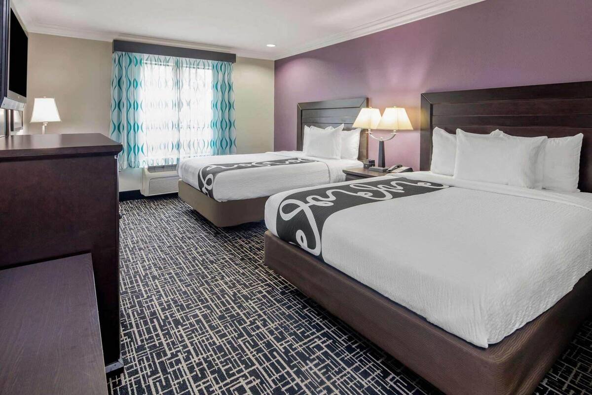 A hotel room with two beds, a desk, a TV, and a window with curtains.