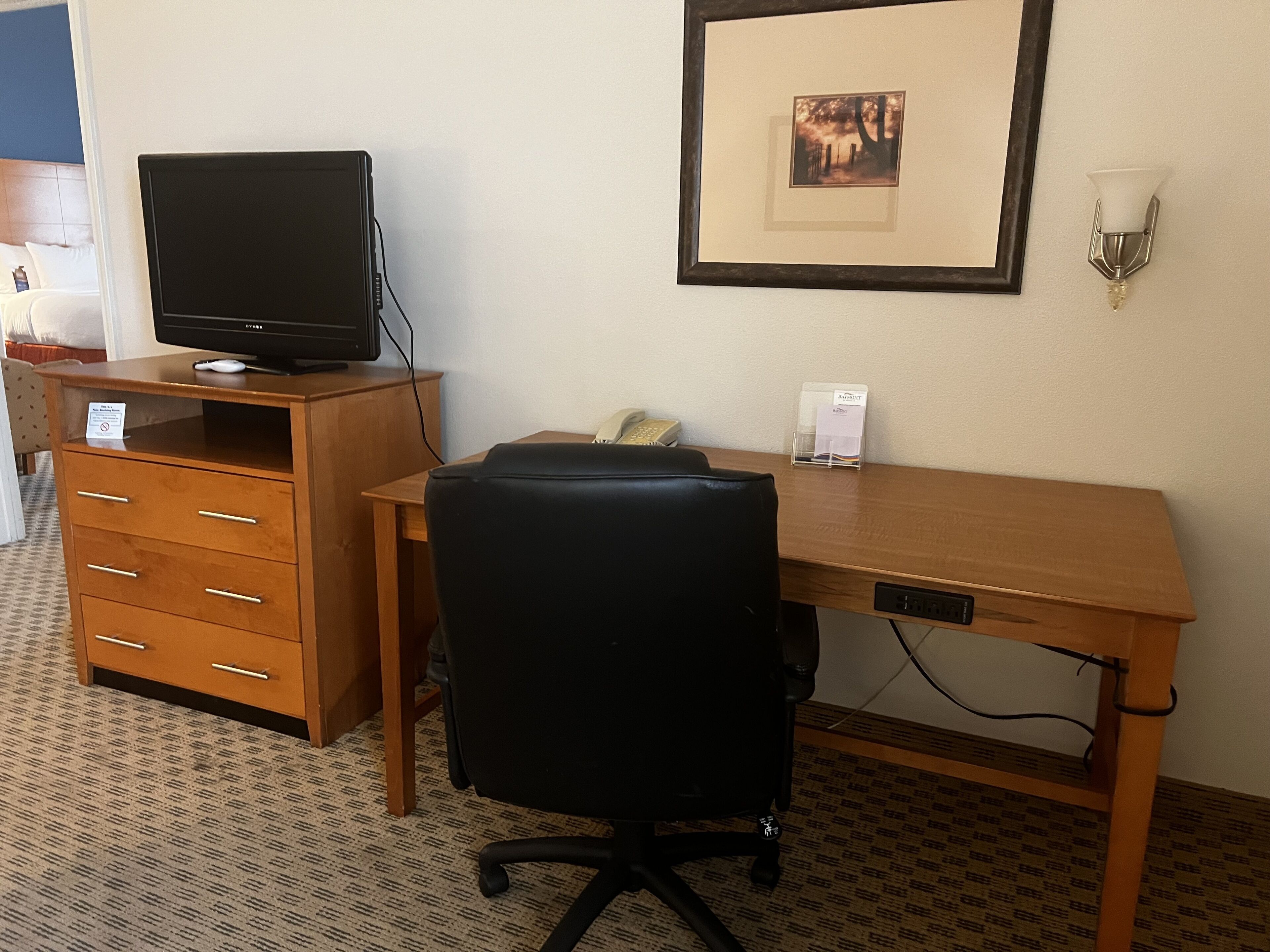 A hotel room with a wooden desk, a black office chair, a television on a wooden cabinet, a framed picture on the wall, and a bedside table with a lamp.