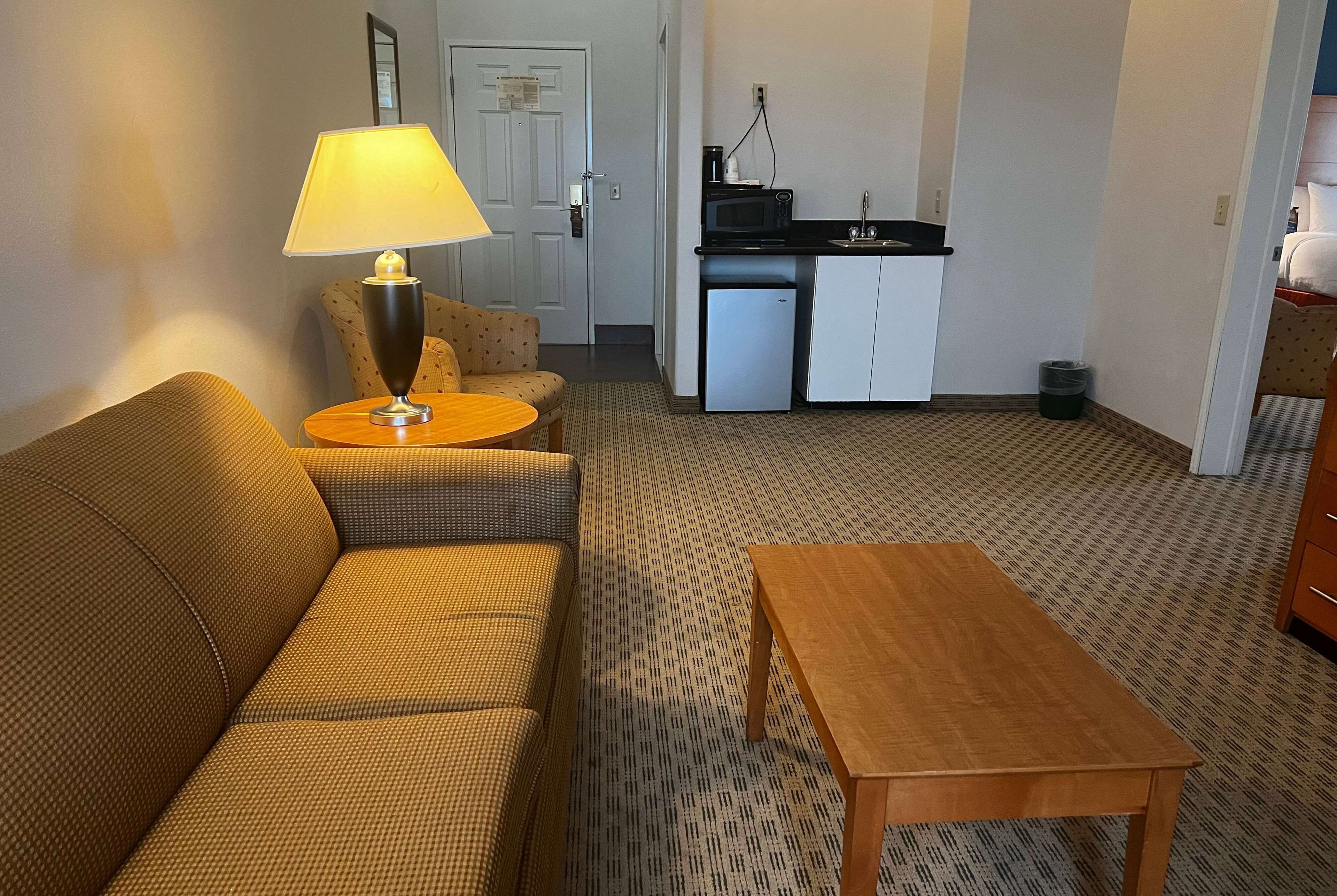 En kompakt opholdsstue med sofa, rundt bord med lampe, lille tekøkken og et gulvtæppe med et mønster.