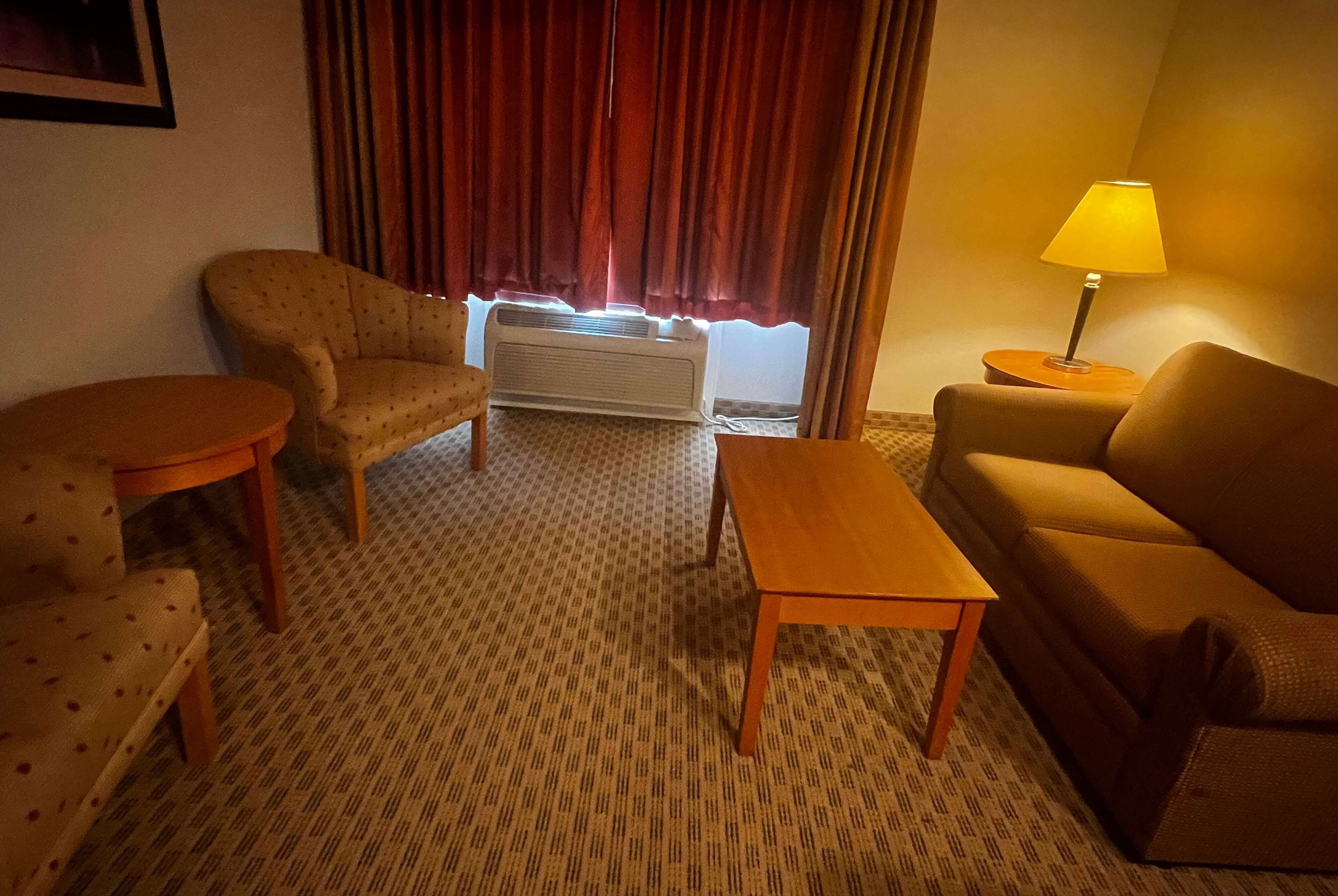 Una habitación de hotel con alfombra estampada, un sofá, una silla, una mesita, una lámpara y una ventana con cortinas.