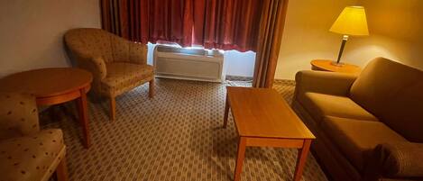 A hotel room with a patterned carpet, a sofa, a chair, a small table, a lamp, and a window with curtains.