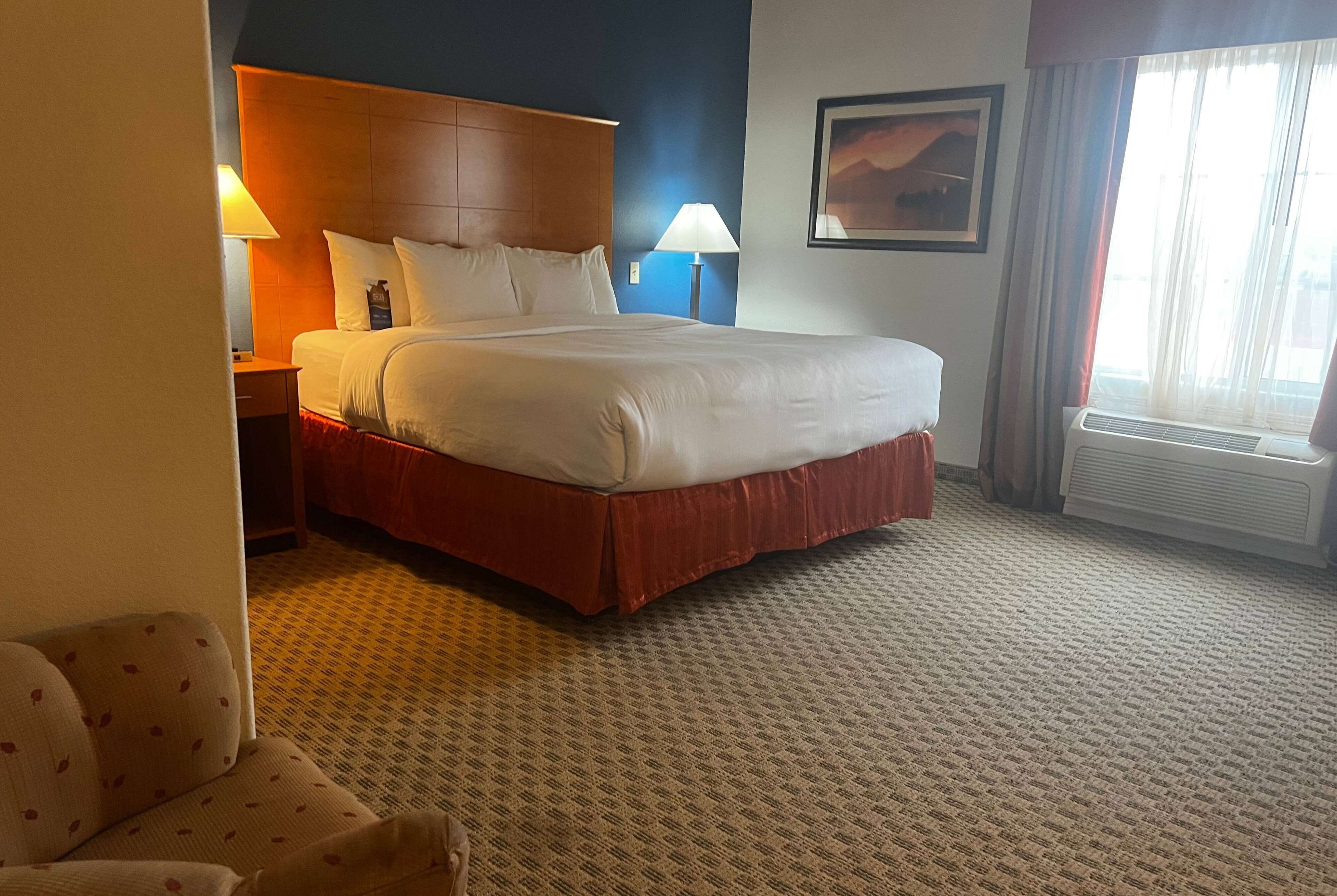 Una habitación de hotel con una cama grande, dos mesitas de noche, una silla, un cuadro en la pared y una ventana con cortinas.