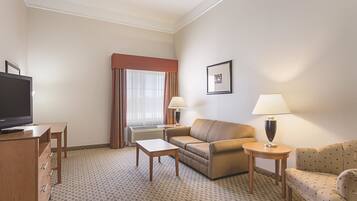 A hotel room with a flat-screen TV, two couches, a coffee table, two lamps, and a large window with curtains.