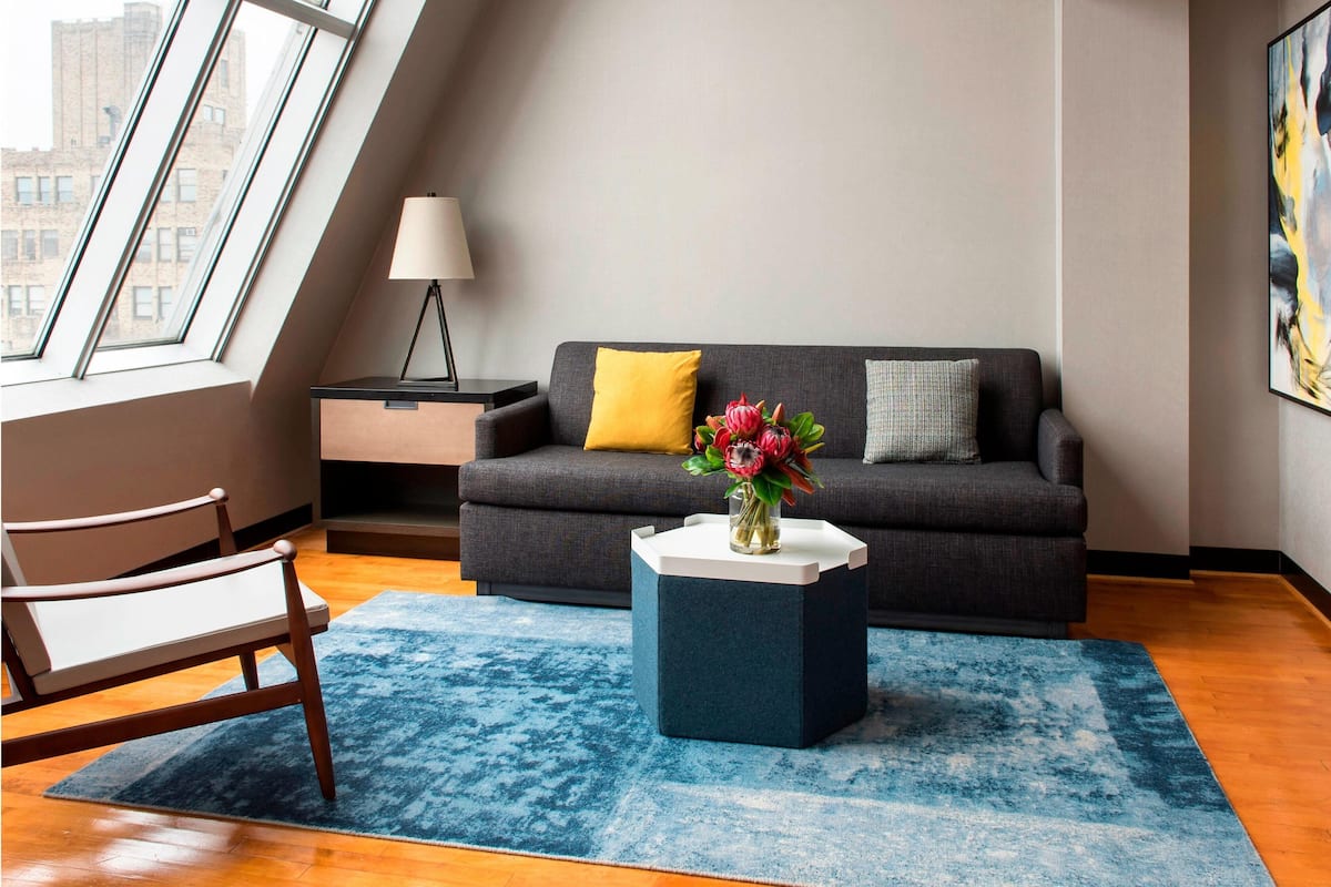 Un salon moderne avec un canapé gris foncé, un tapis bleu, une chaise en bois, une petite table sur laquelle est posé un vase de fleurs et une lampe sur une table d’appoint.