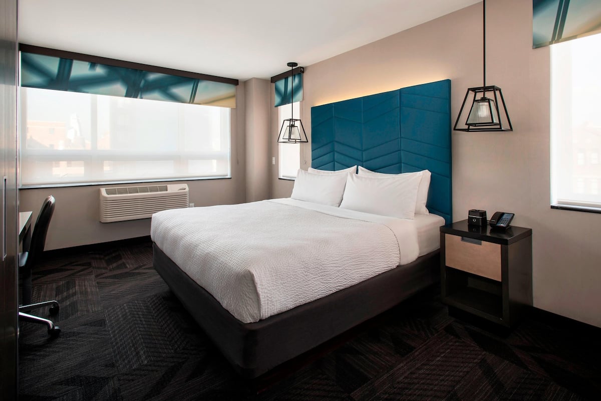 Une chambre d’hôtel moderne, dotée d’un grand lit, d’un bureau et d’une chaise. On y trouve une fenêtre avec des stores, une table de chevet avec une lampe et un climatiseur fixé au mur.