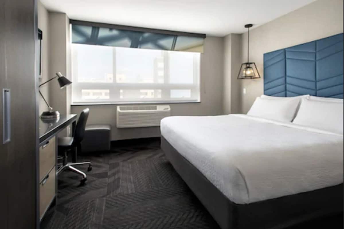 Une chambre d’hôtel moderne dotée d’un grand lit, d’un bureau avec une chaise, d’une fenêtre avec des stores et d’un climatiseur fixé au mur.