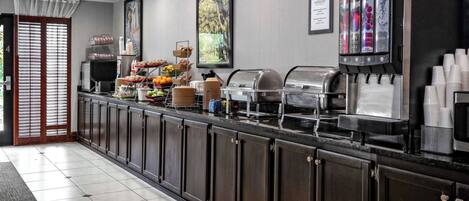 A hotel breakfast area with a buffet setup, including a coffee machine, fruit display, and various food items.