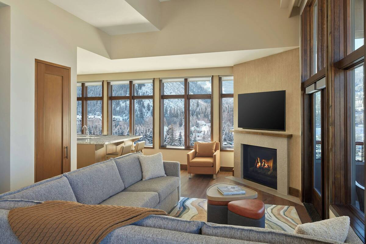 A spacious living room with a large sectional sofa, a fireplace, a television mounted on the wall, and large windows offering a view of a snowy landscape.