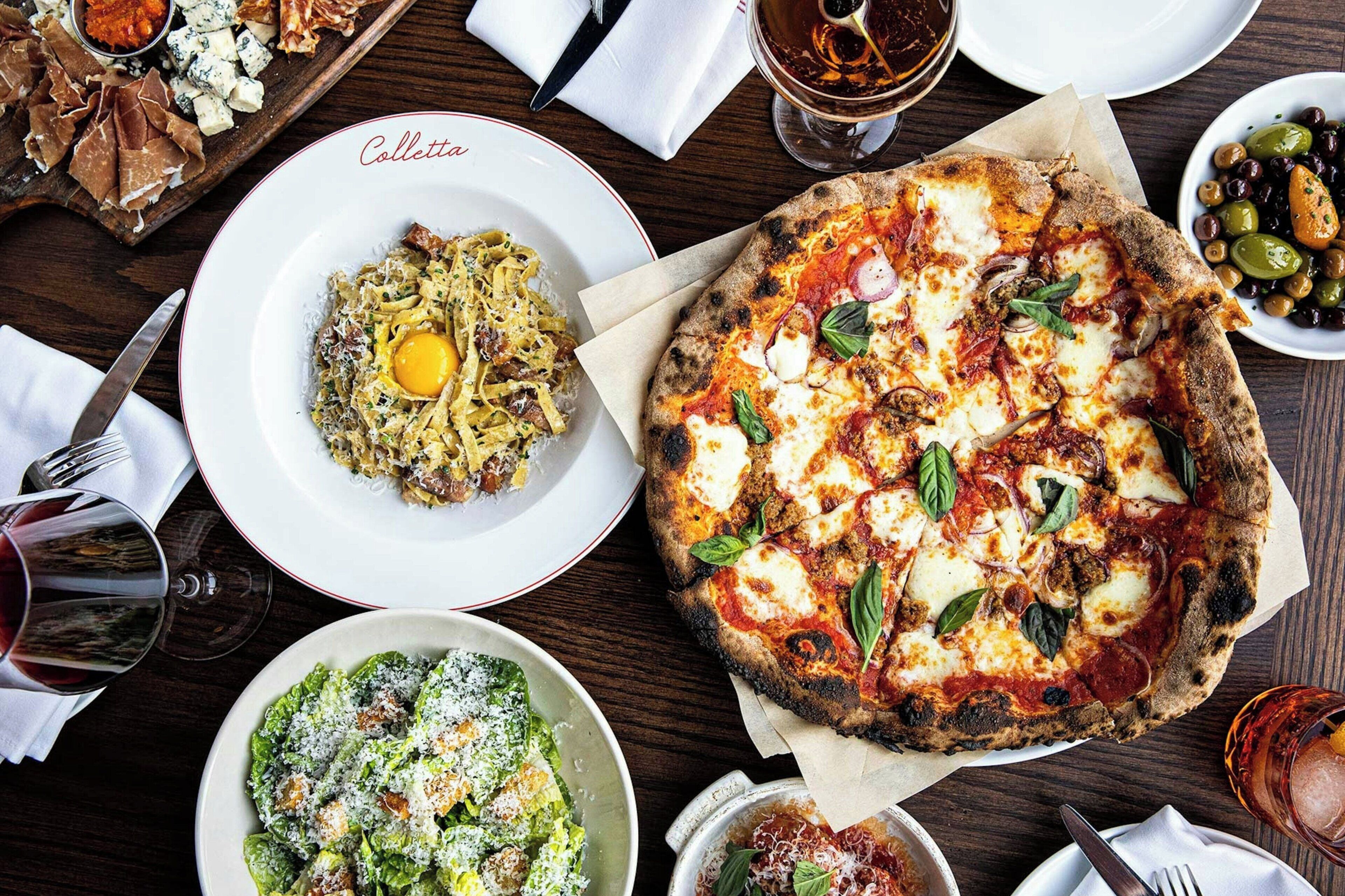 Une table garnie de différents plats, notamment des pâtes, une pizza et une salade, ainsi que des verres contenant une boisson sombre.