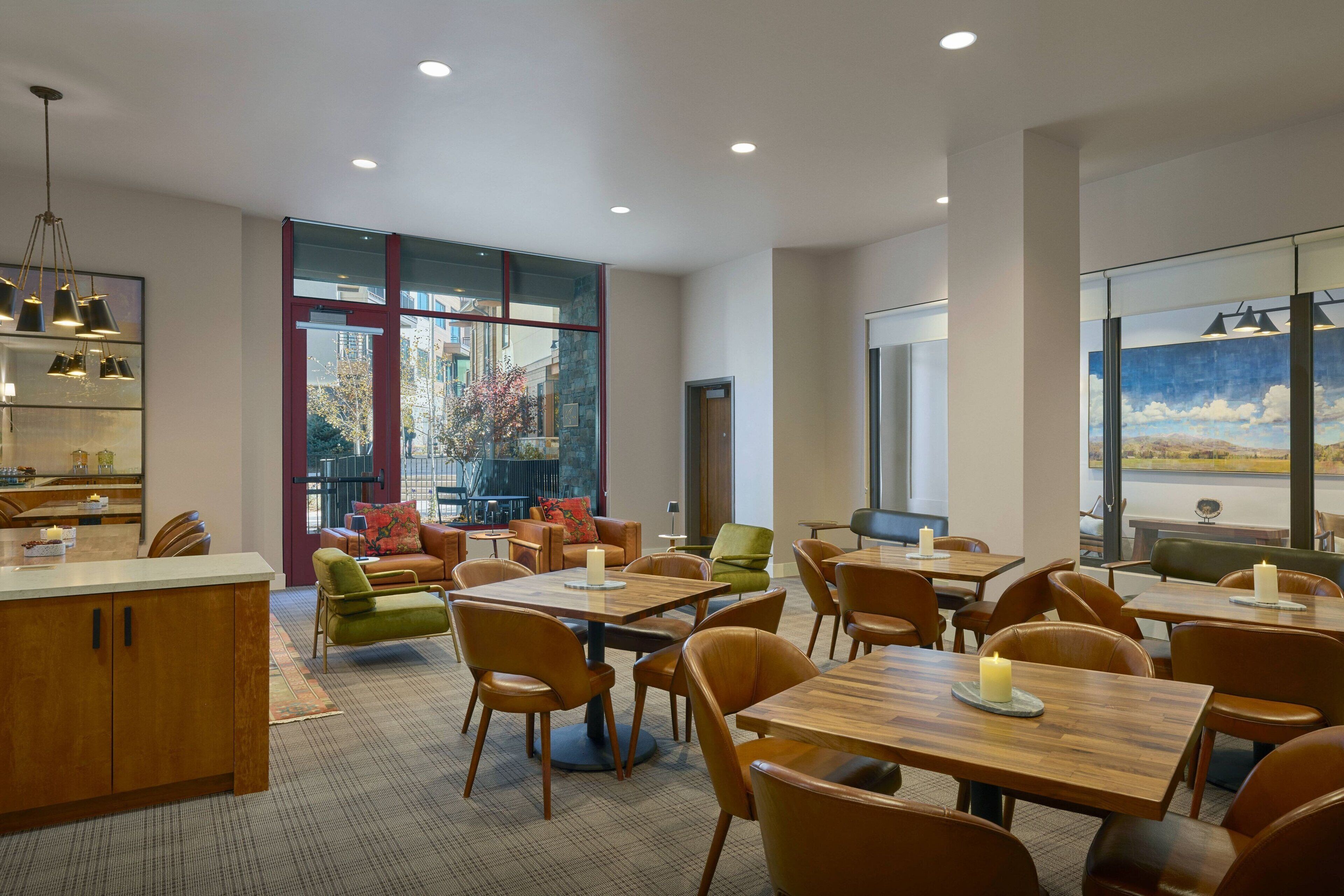 A modern dining area with wooden tables and chairs, large windows with a view of buildings, and a mix of seating options including a sofa and chairs.