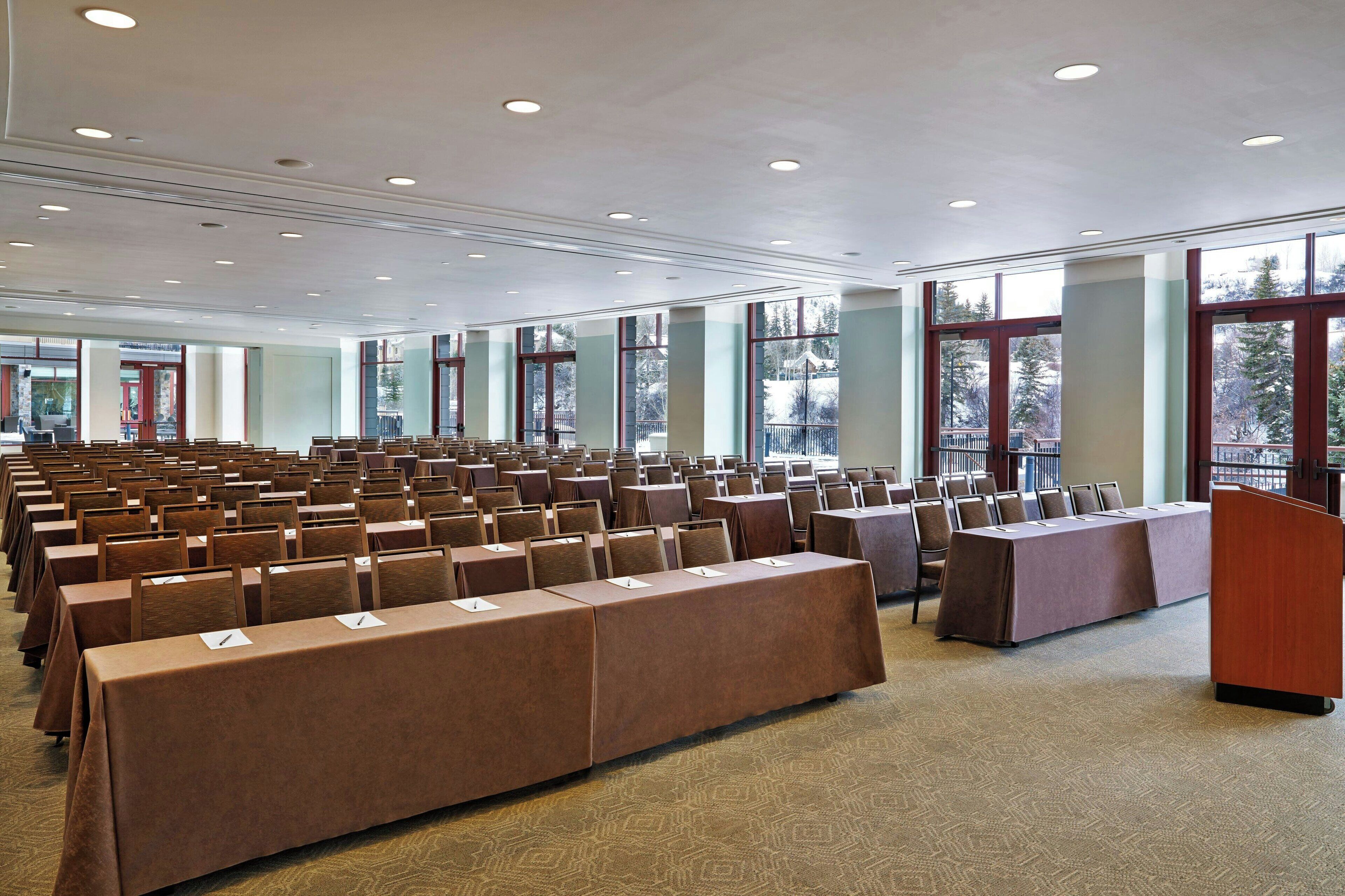 Une salle de conférence avec des rangées de tables et de chaises, de grandes fenêtres et un pupitre.