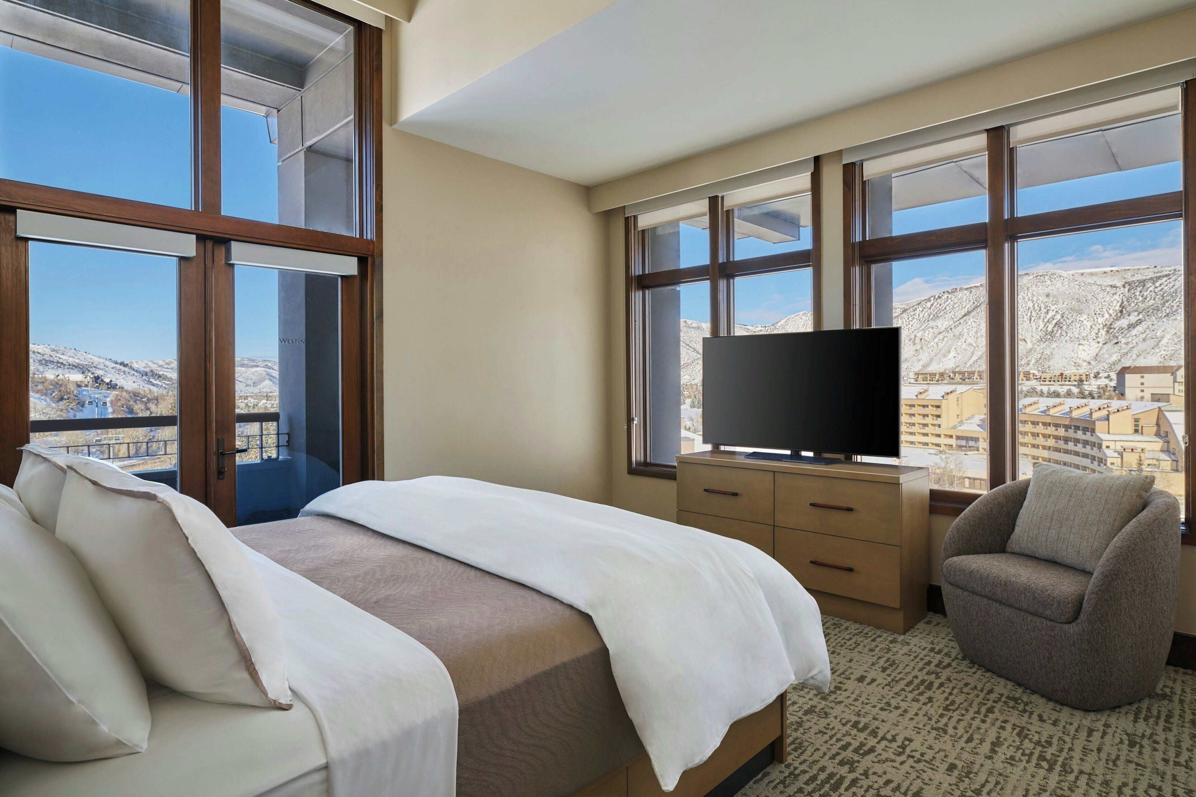 A hotel room with a large bed, a TV on a wooden dresser, a chair, and large windows offering a mountain view.