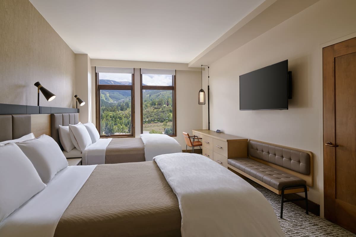 A hotel room with two beds, a desk, a bench, a TV, and a large window with a view of the outdoors.