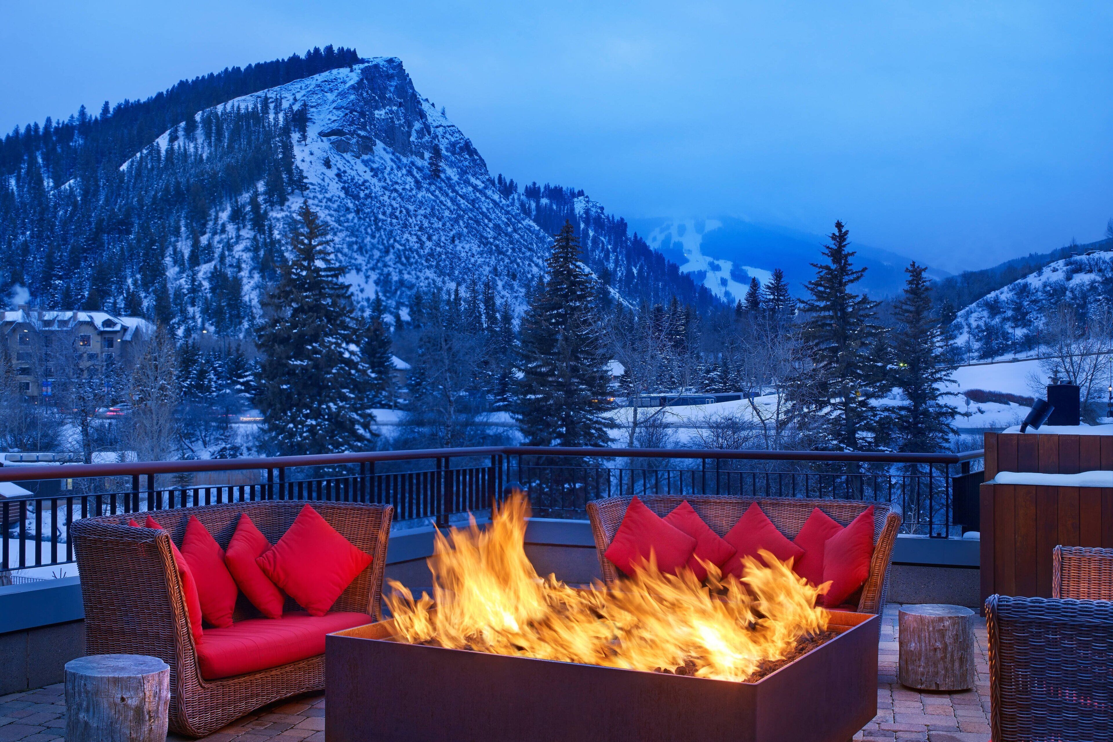 A fire pit area with wicker furniture and red cushions, overlooking a snowy mountain landscape.