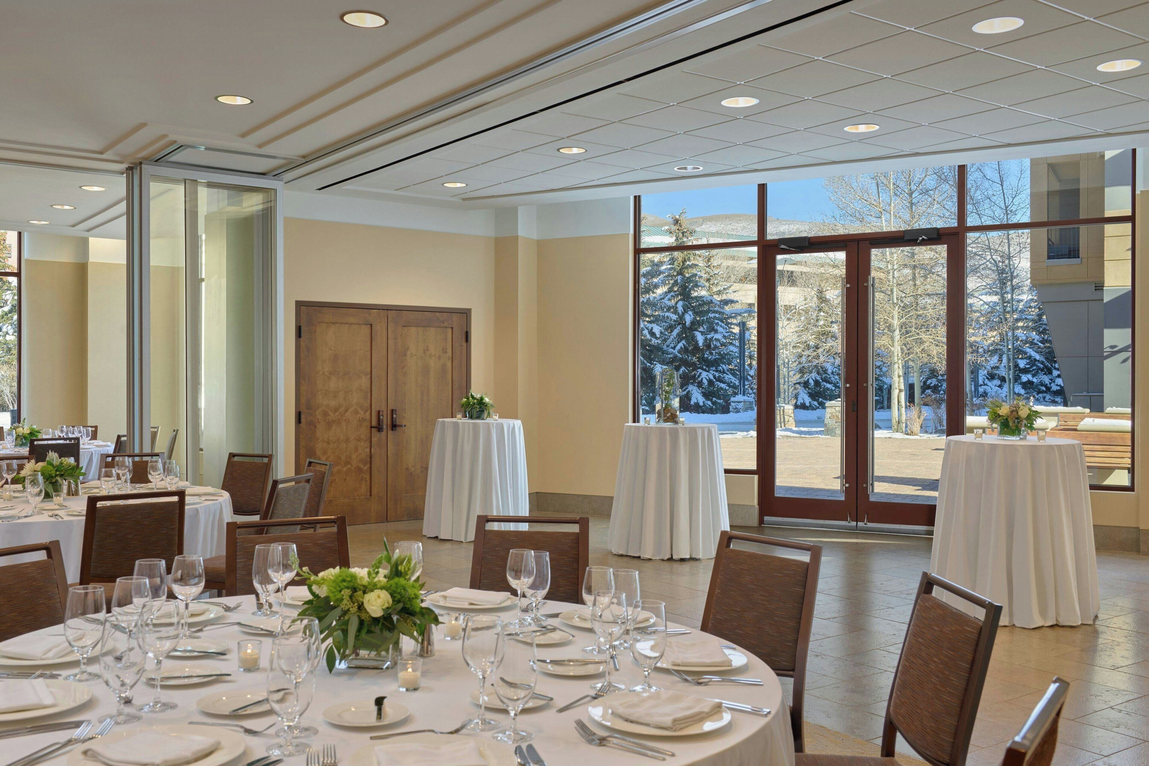 A dining area with round tables set for a meal, large windows offering a view of snow-covered trees, and a door leading to another room.