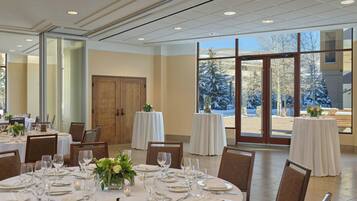 A dining area with round tables set for a meal, large windows offering a view of snow-covered trees, and a door leading to another room.