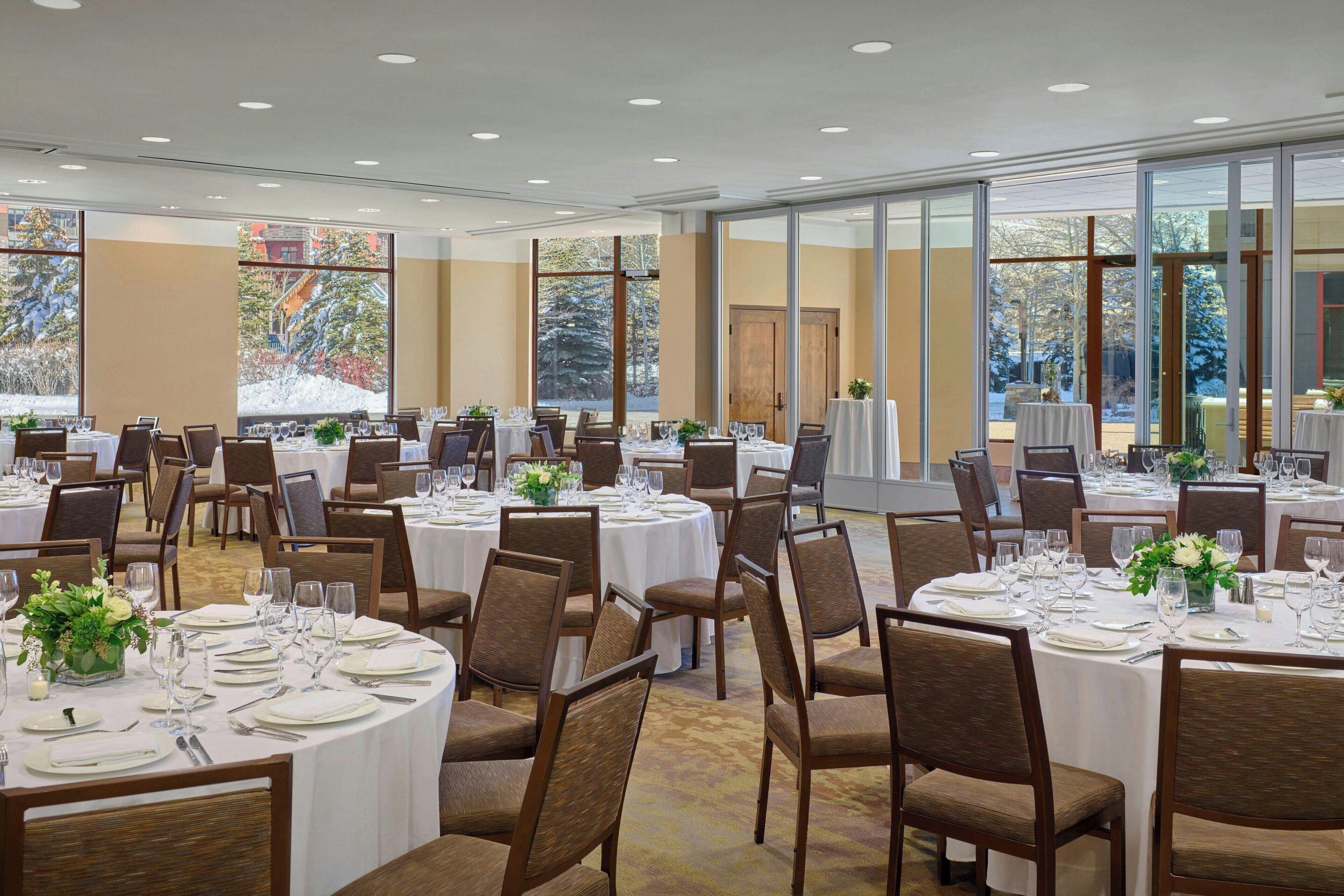 A banquet hall with round tables set for dining, large windows offering a view of trees and a snowy landscape.