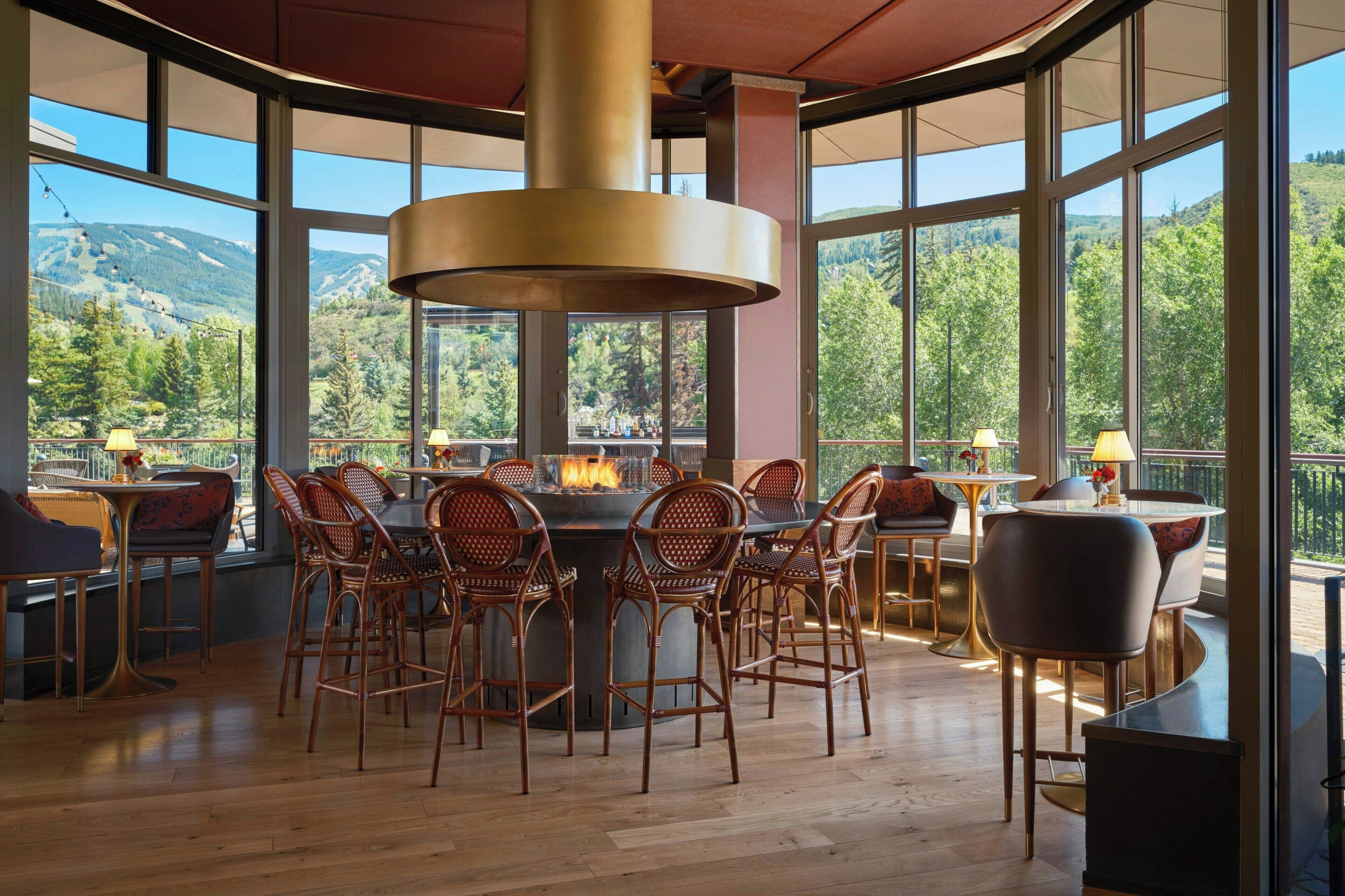 A dining area with a central fire feature, surrounded by wicker chairs and tables, with a view of a mountainous landscape through large windows.
