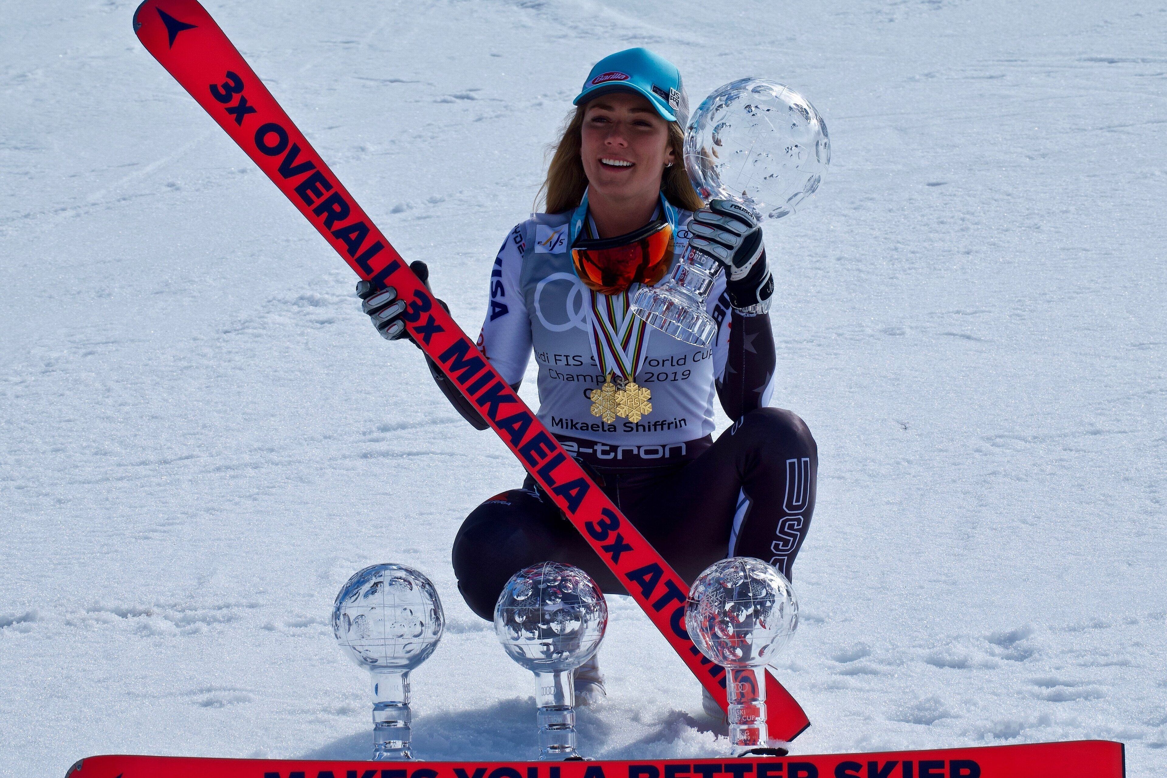 Una persona sosteniendo un trofeo y un esquí con la inscripción '3x OVERALL'.