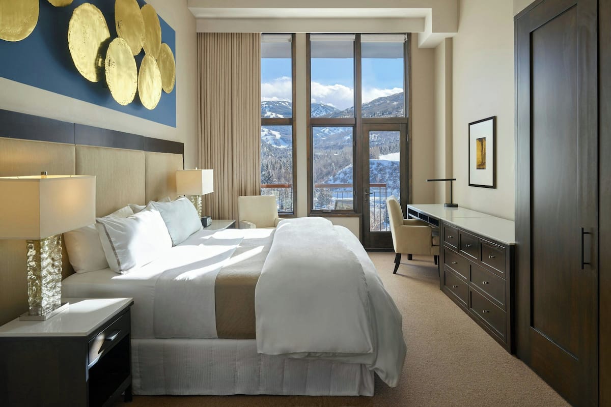 A hotel room with a large bed, a desk, a chair, and a view of mountains through large windows.