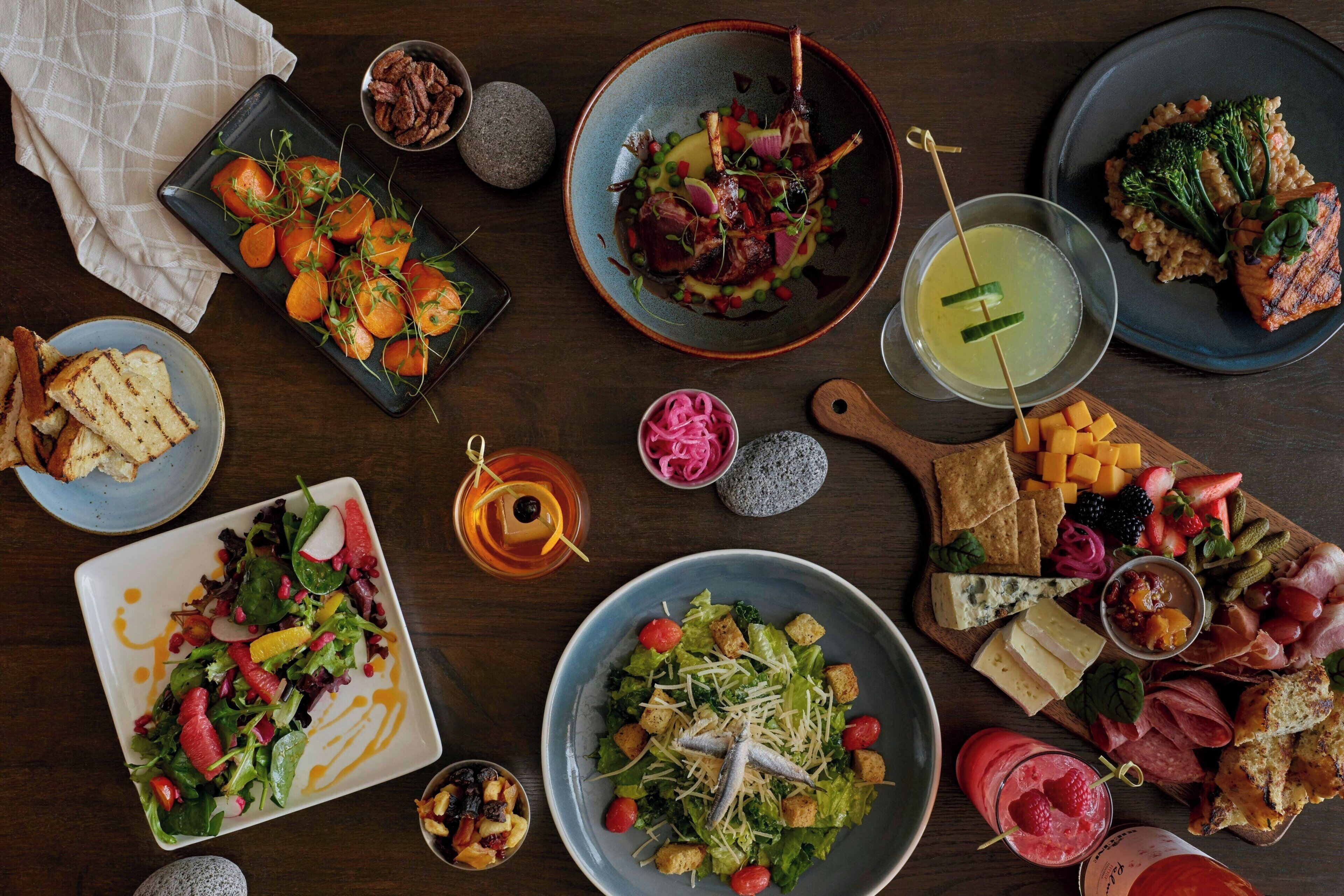 A variety of dishes and drinks on a wooden table, including salads, grilled meats, and cheese.